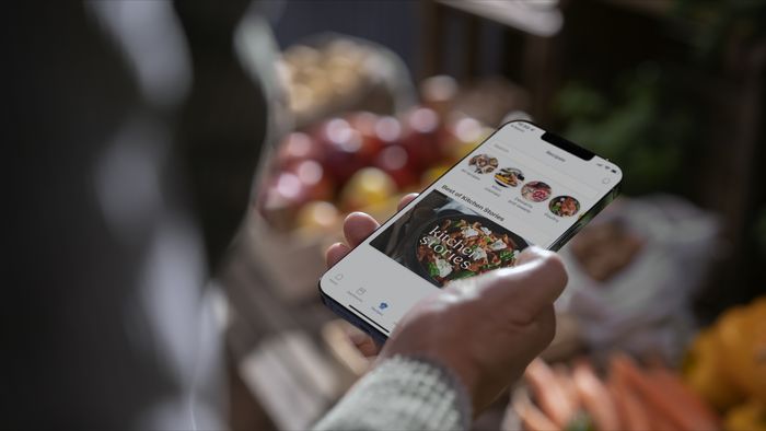 Persona utilizando un móvil en un mercado, mirando recetas o ideas culinarias a través de la aplicación Home Connect que destaca 'Kitchen Stories', con frutas frescas visibles en el fondo. 